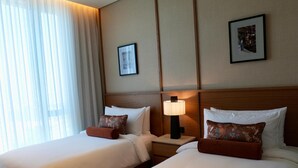Habitación de hotel con dos camas, una mesita de noche con lámpara y un ventanal con cortinas traslúcidas.