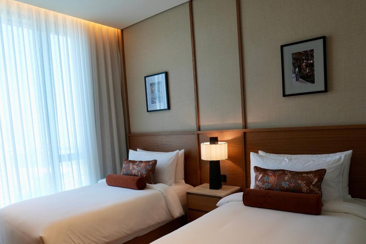 A hotel room with two beds, a nightstand with a lamp, and a large window with sheer curtains.