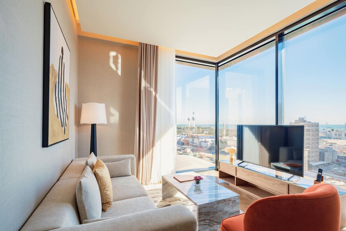 A modern hotel room with a large window, a sofa, a red chair, a TV, and a lamp.