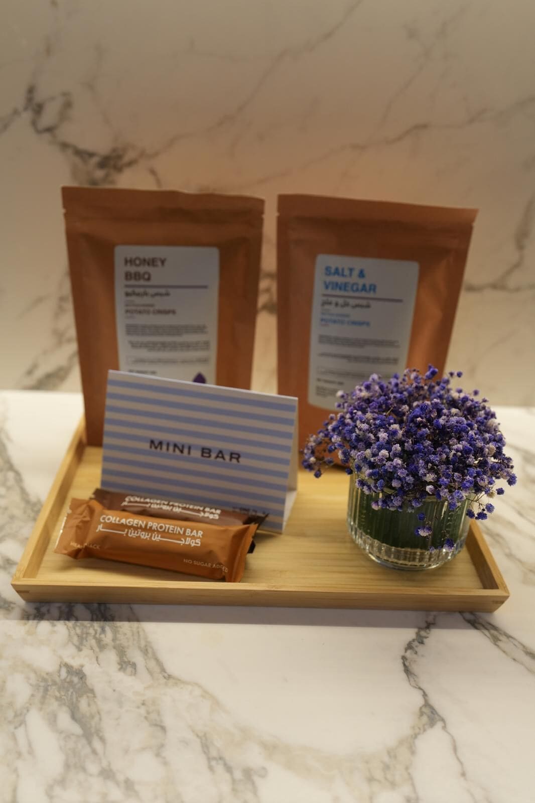 A tray with a mini bar setup including a honey BBQ packet, a salt & vinegar packet, a collagen protein bar, and a vase with purple flowers.