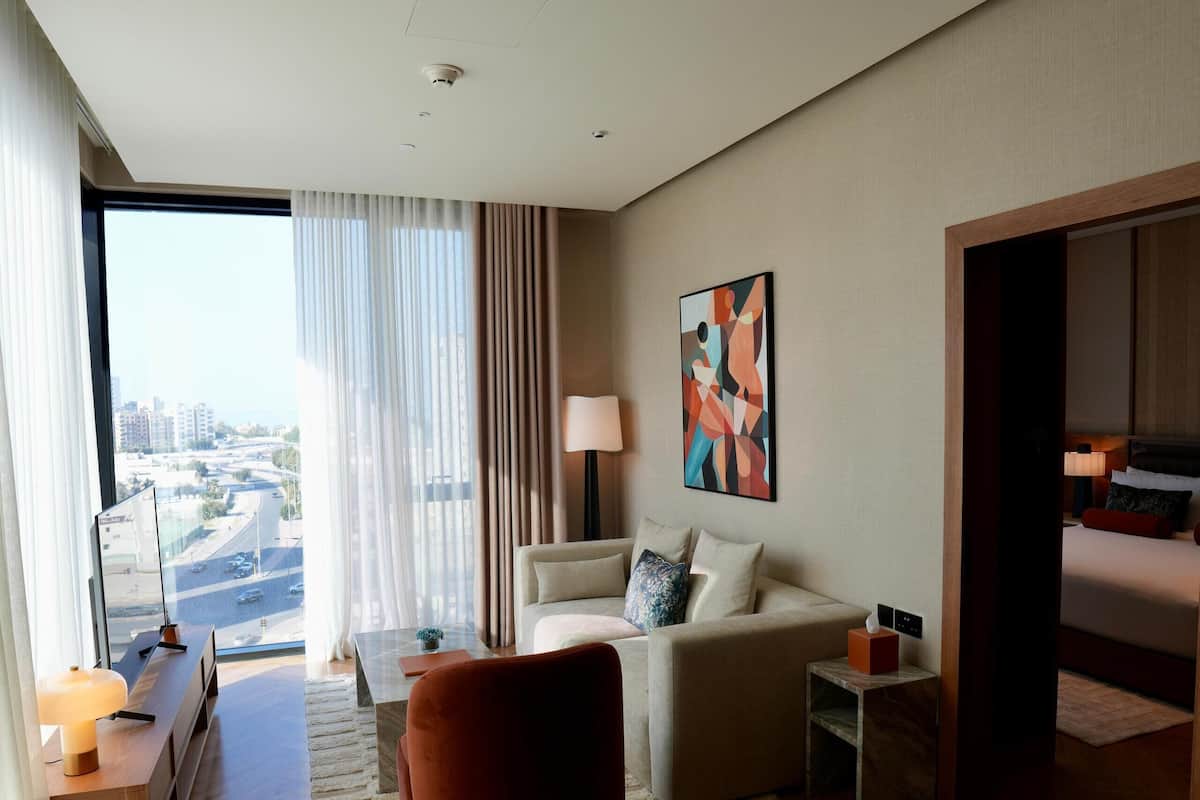 A modern hotel room with a large window, a sofa, a coffee table, and a red ottoman.