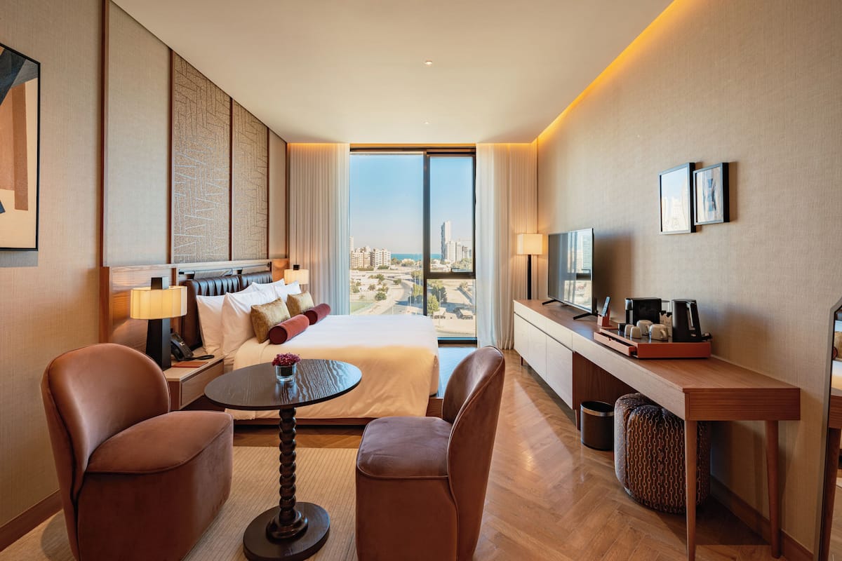 A hotel room with a large bed, two armchairs, a small round table, a desk with a TV, and a view of the cityscape.
