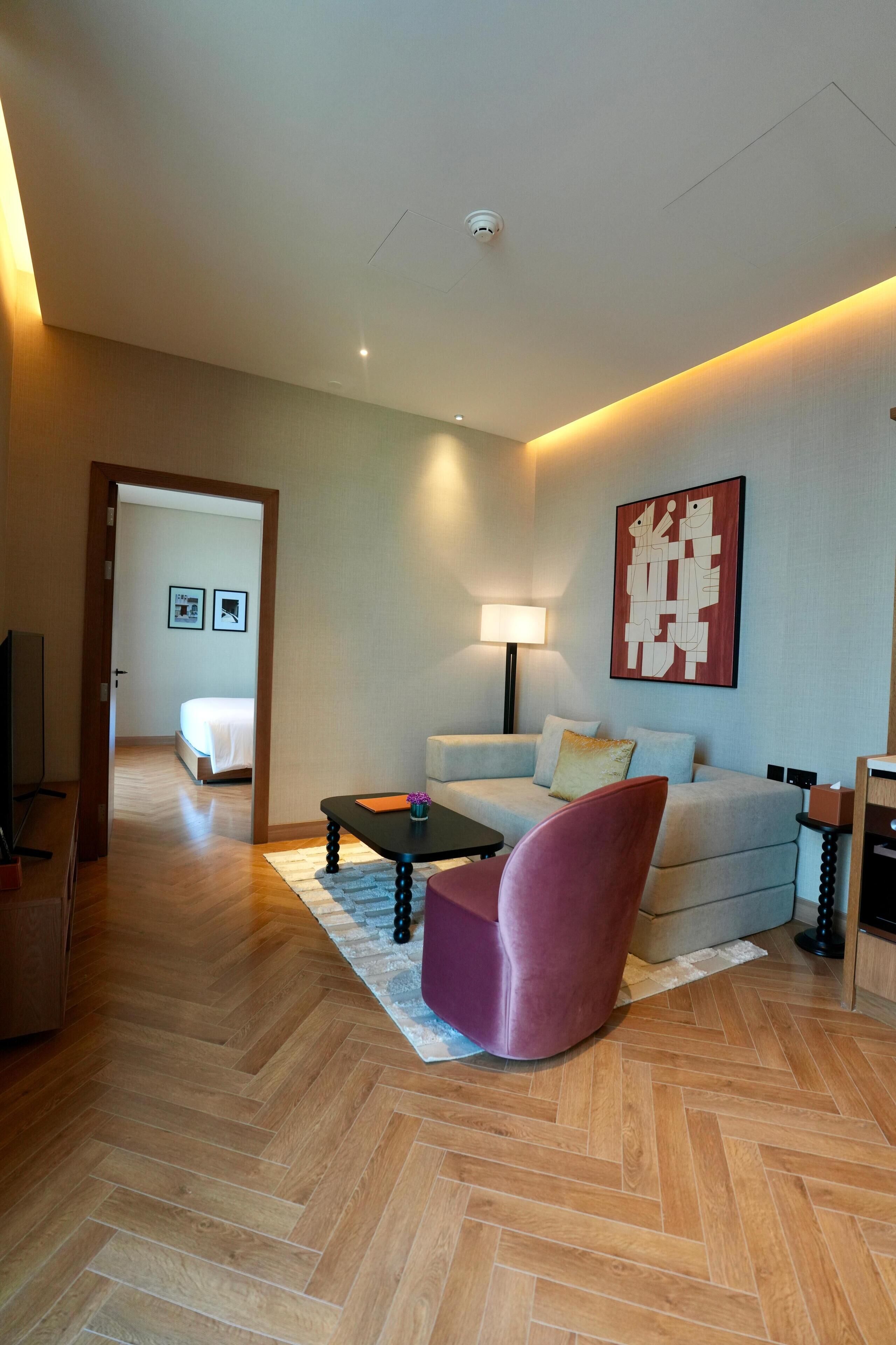 A modern hotel room with a wooden herringbone floor, a sofa, a chair, a small table, a lamp, and a painting on the wall.