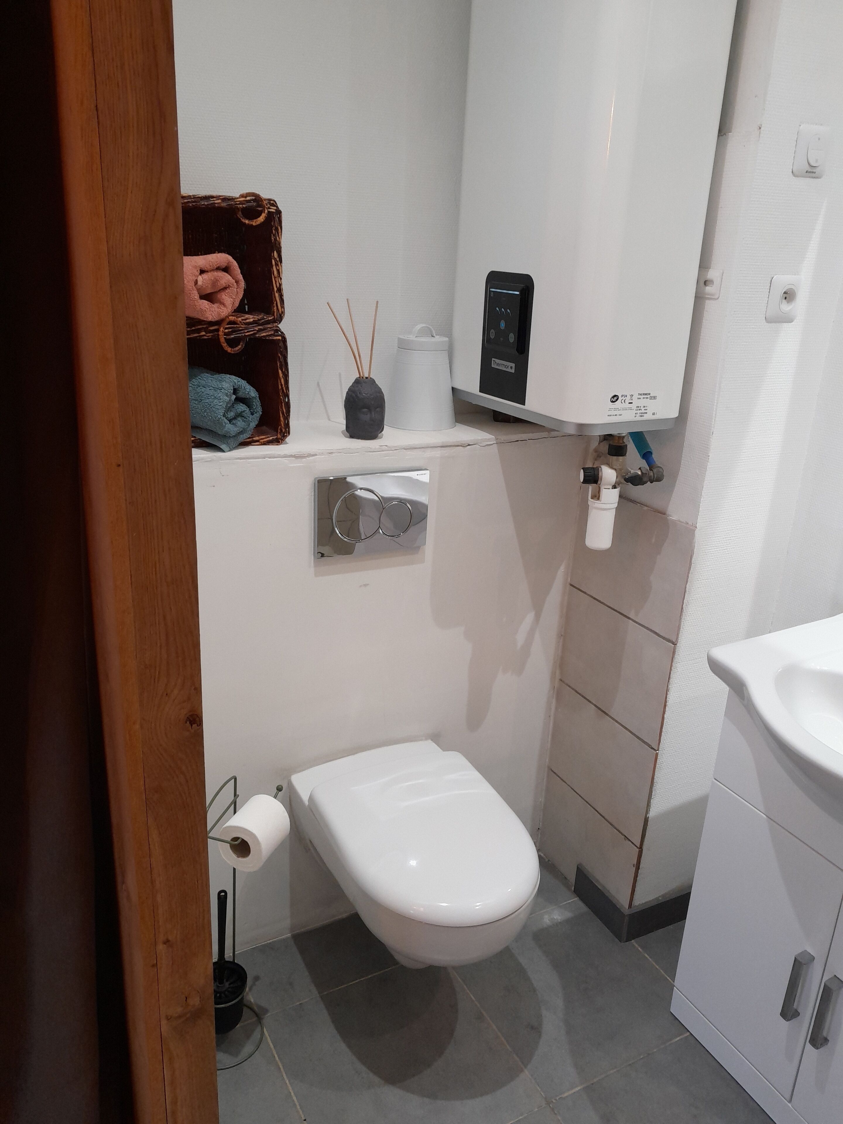 Un bany petit i funcional, amb un vàter blanc, un calefactor penjat a la paret, una petita prestatgeria amb un ram i una tovallola, i un suport de paper higiènic.