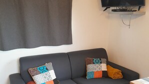A dark grey sofa with patterned cushions and a rolled-up towel, a flat-screen TV mounted on the wall, and a small wooden table with a white surface. - A cosy little nest (Droisy)