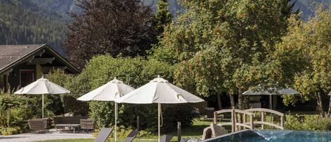 A swimming pool with lounge chairs and umbrellas, surrounded by trees and mountains in the background.