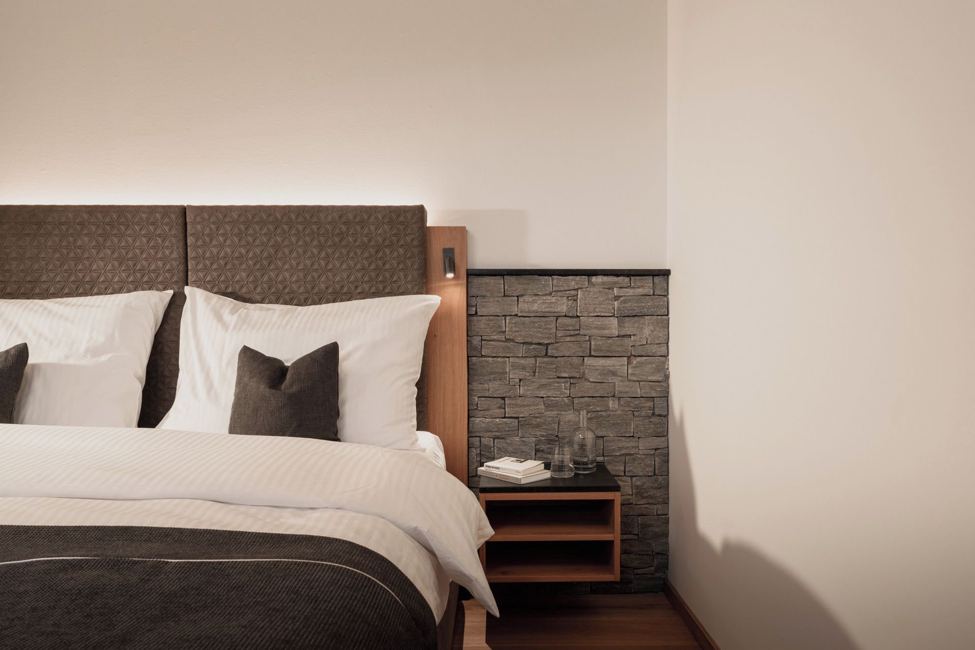 A neatly made bed with a headboard, a nightstand with a stone wall, and a small shelf.