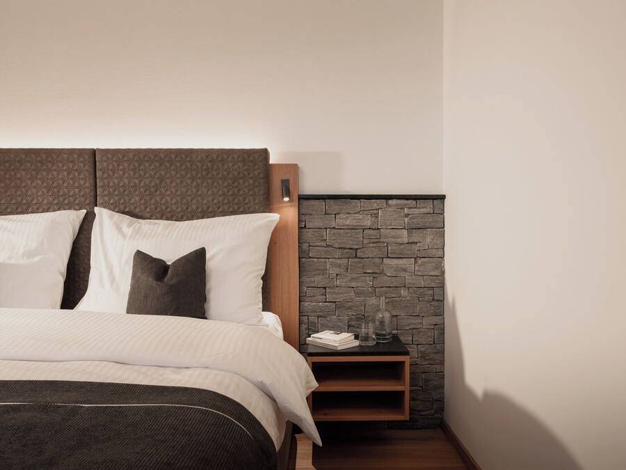 Una cama bien hecha con cabecera, mesita de noche y un pequeño estante al lado de una pared de piedra.