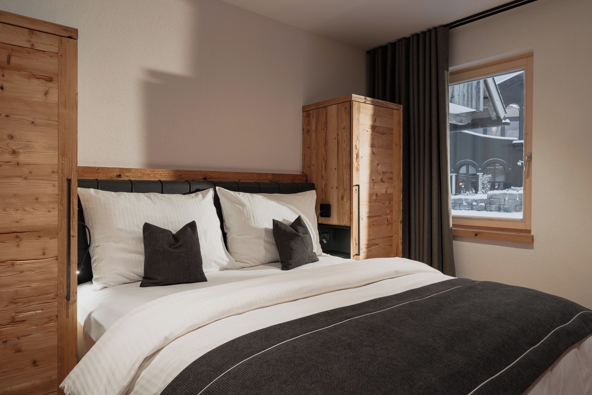 Ein Schlafzimmer mit einem hölzernen Kopfende, einem Bett mit weißen und dunklen Kissen, einem Holzschrank und einem Fenster mit Blick auf eine verschneite Landschaft.