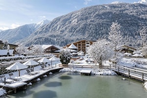 Un espacio al aire libre cubierto de nieve, con sillones de jardín y sombrillas, un puente de madera y edificios al fondo.