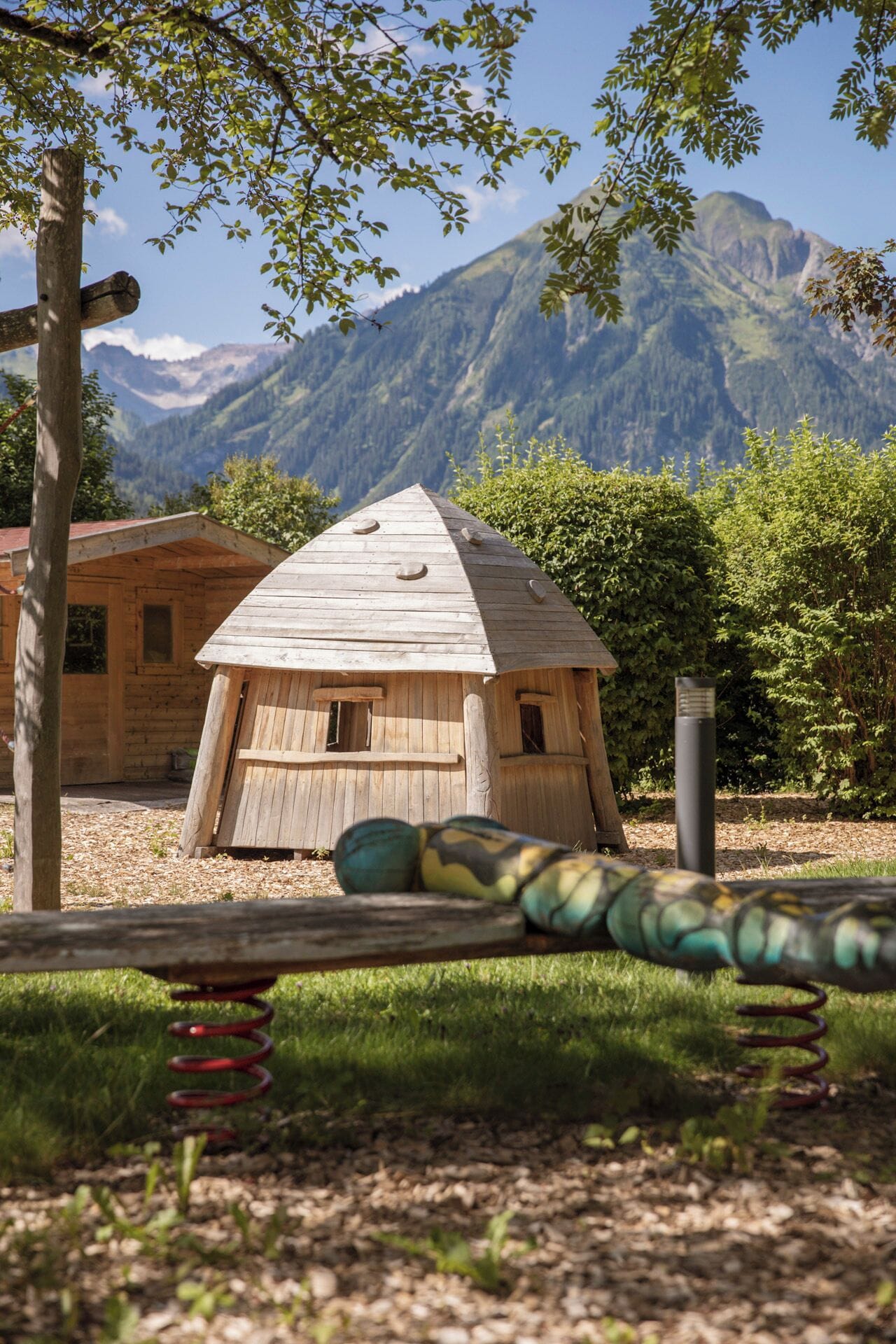 A wooden structure with a sloped roof, surrounded by greenery and mountains in the background.