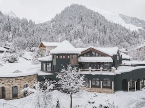 Un edificio grande de varios pisos con un balcón destacado, rodeado de árboles nevados y montañas.