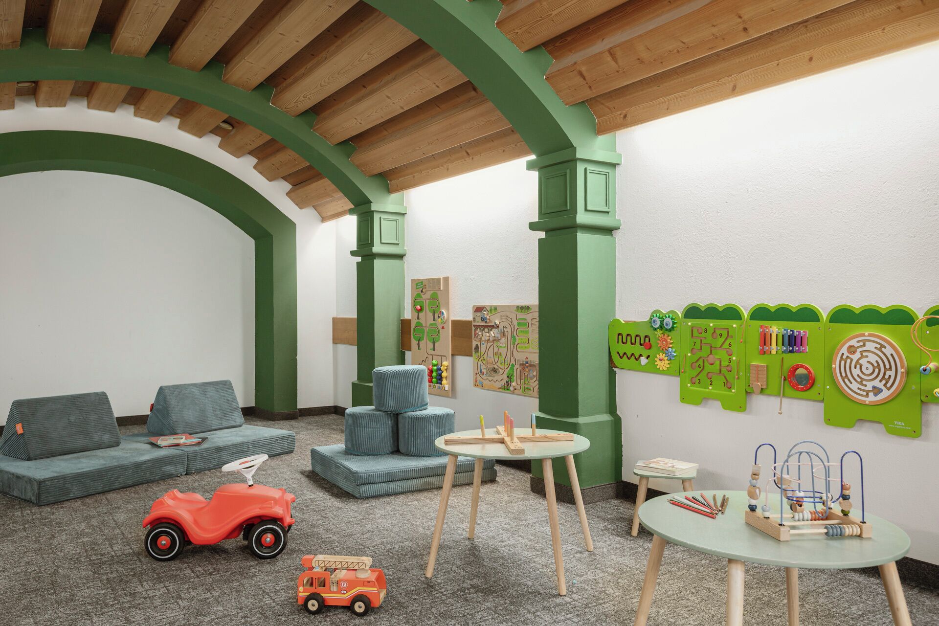 Ein Kinderspielzimmer mit einem grünen Bogen, gepolsterten Sitzgelegenheiten, lehrreichen Spielzeugen und einer Holzbodenverkleidung.
