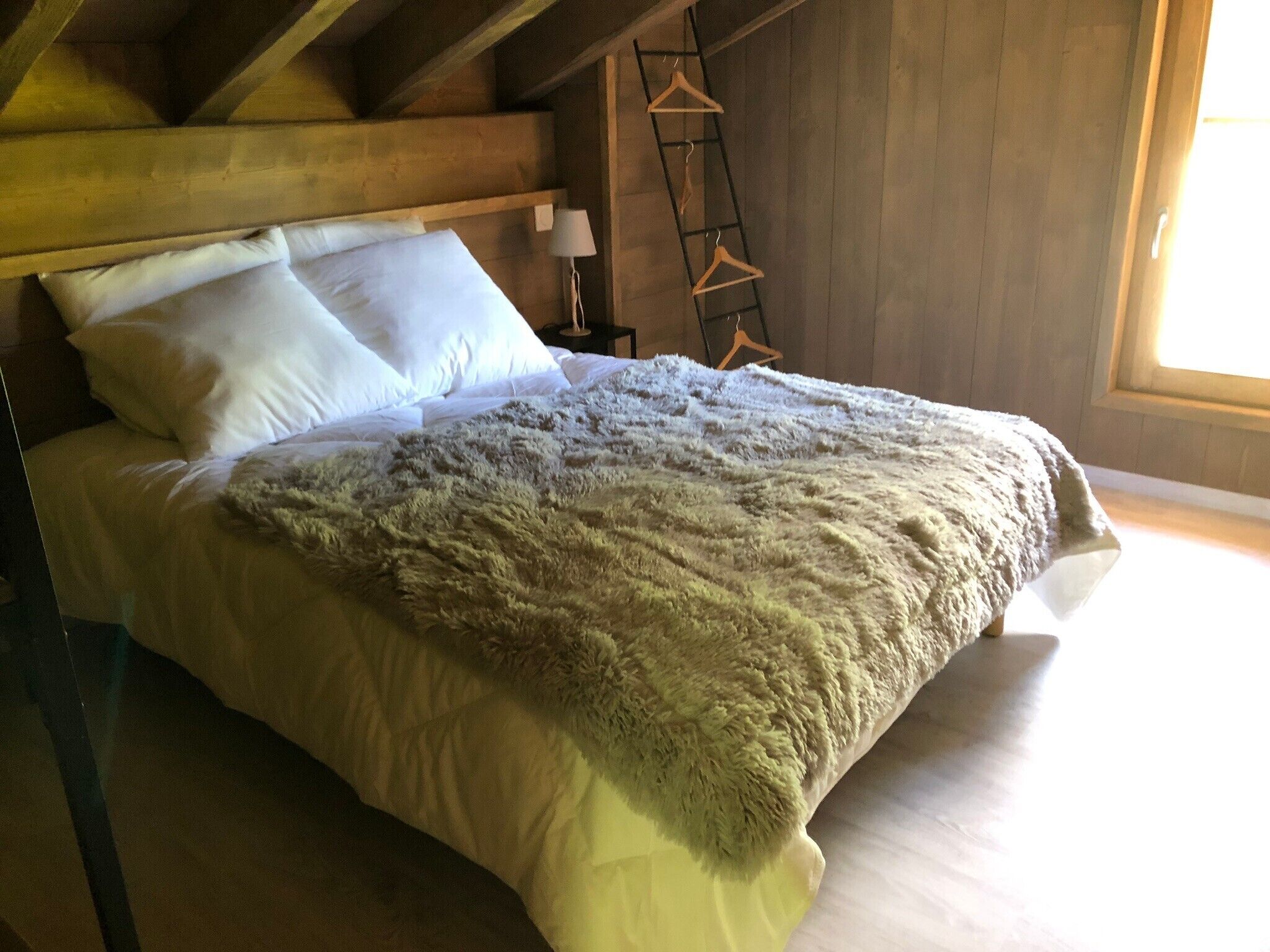 A bed with white linens and a fluffy blanket, wooden beams above, a ladder, and a hanging rack against the wall.