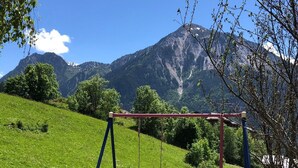 Une aire de jeux avec des balançoires et un espace délimité par une clôture, entourée d’un champ verdoyant et avec des montagnes en arrière-plan.