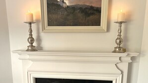Un tableau encadré représentant un paysage avec une maison située sur une colline, et deux bougies allumées de part et d’autre sur une cheminée.