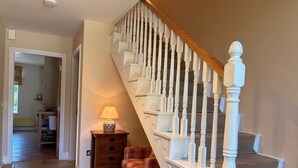 Un escalier avec des balustrades blanches et une main courante en bois mène à l’étage supérieur. Un fauteuil à carreaux est placé sur la plateforme, et une lampe de table se trouve sur une table de chevet.