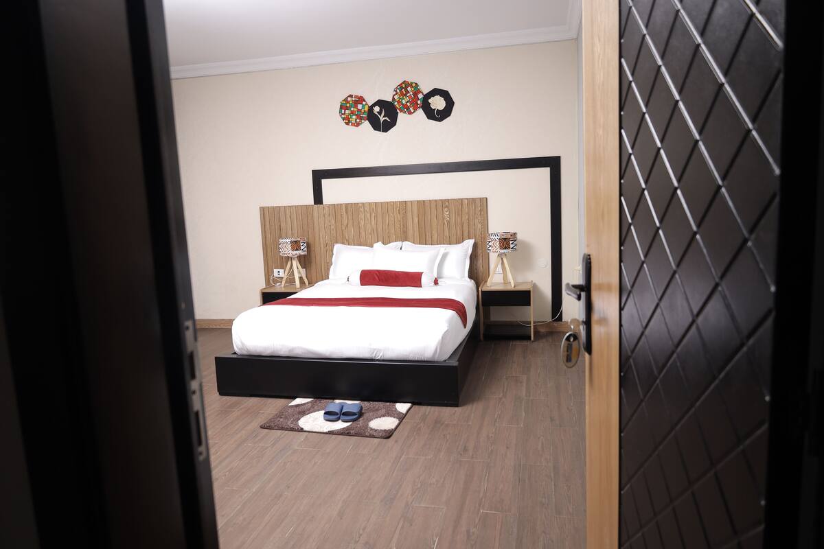 A modern bedroom with a wooden headboard, a bed with white bedding and a red accent pillow, a nightstand with a lamp, and a decorative wall piece.