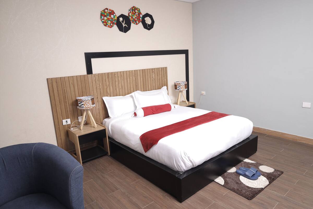 A modern bedroom with a wooden headboard, white bedding, and a red throw. There is a small wooden nightstand with a lamp and a decorative item. A blue chair is visible in the foreground.