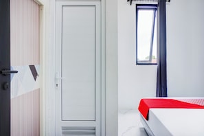A room with a closed door, a window with curtains, and a bed with a red bedspread.
