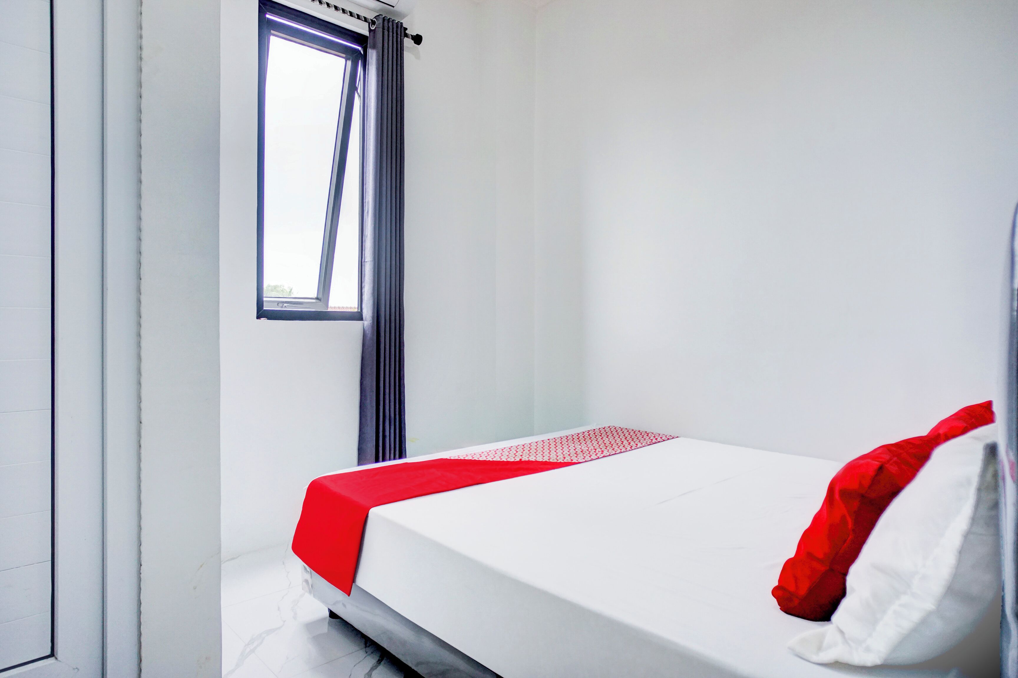 Un lit simple avec un matelas blanc et une literie rouge, dans une pièce aux murs blancs et dotée d’une fenêtre ornée d’un rideau foncé.
