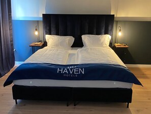 Una cama doble con cabecera oscura, dos mesitas de noche con lámparas y una colcha azul con el texto “HAVEN HOTELS”.