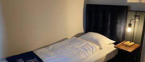 A single bed with a white mattress and a dark headboard. A small bedside table with a lamp is visible. A blue towel with "HAVEN HOTELS" text is draped over the bed.