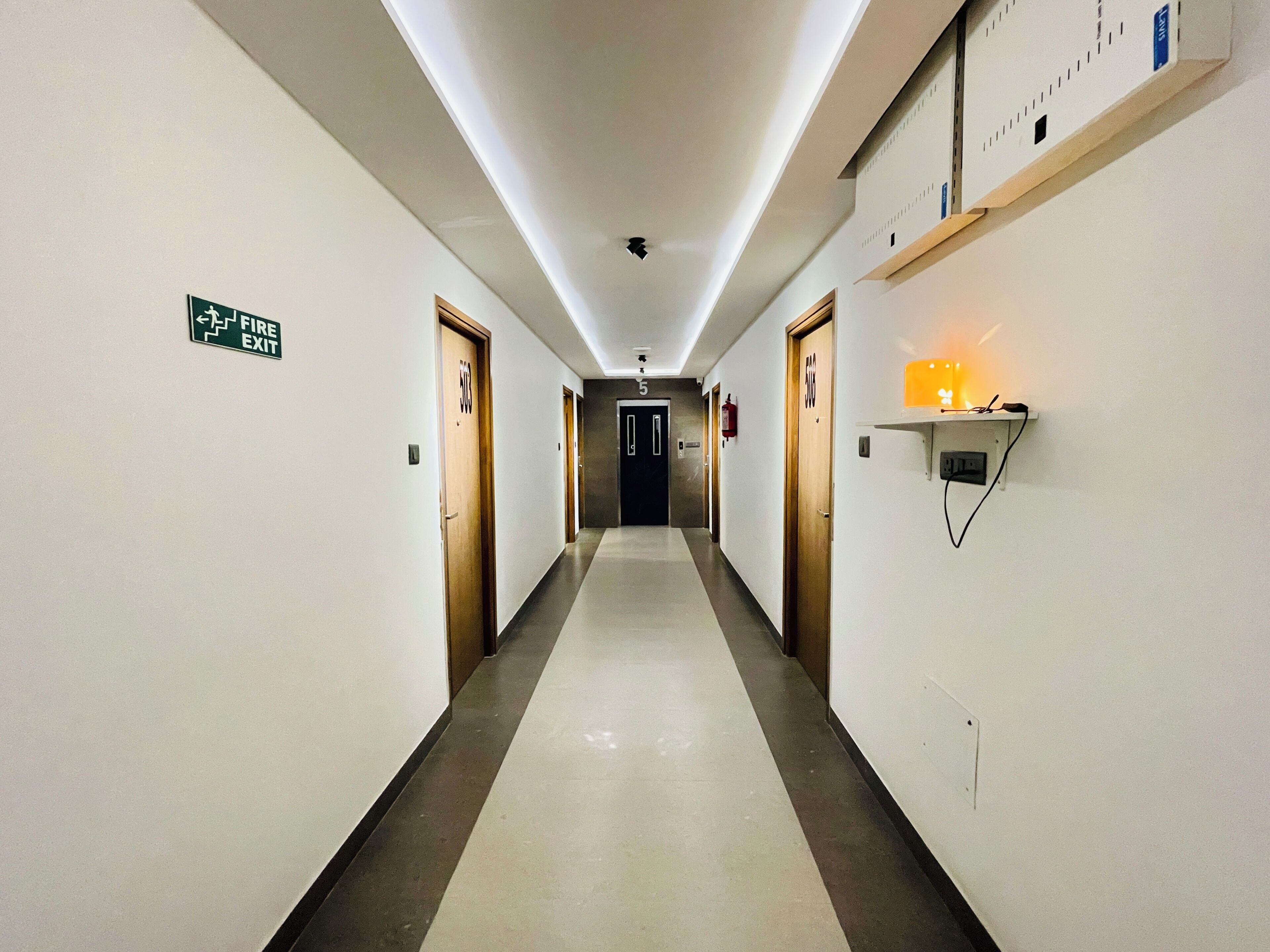 Een hotelgang met een vluchtroutebord, deuren met de nummers 503 en 506, en een wandplank met een lampje.