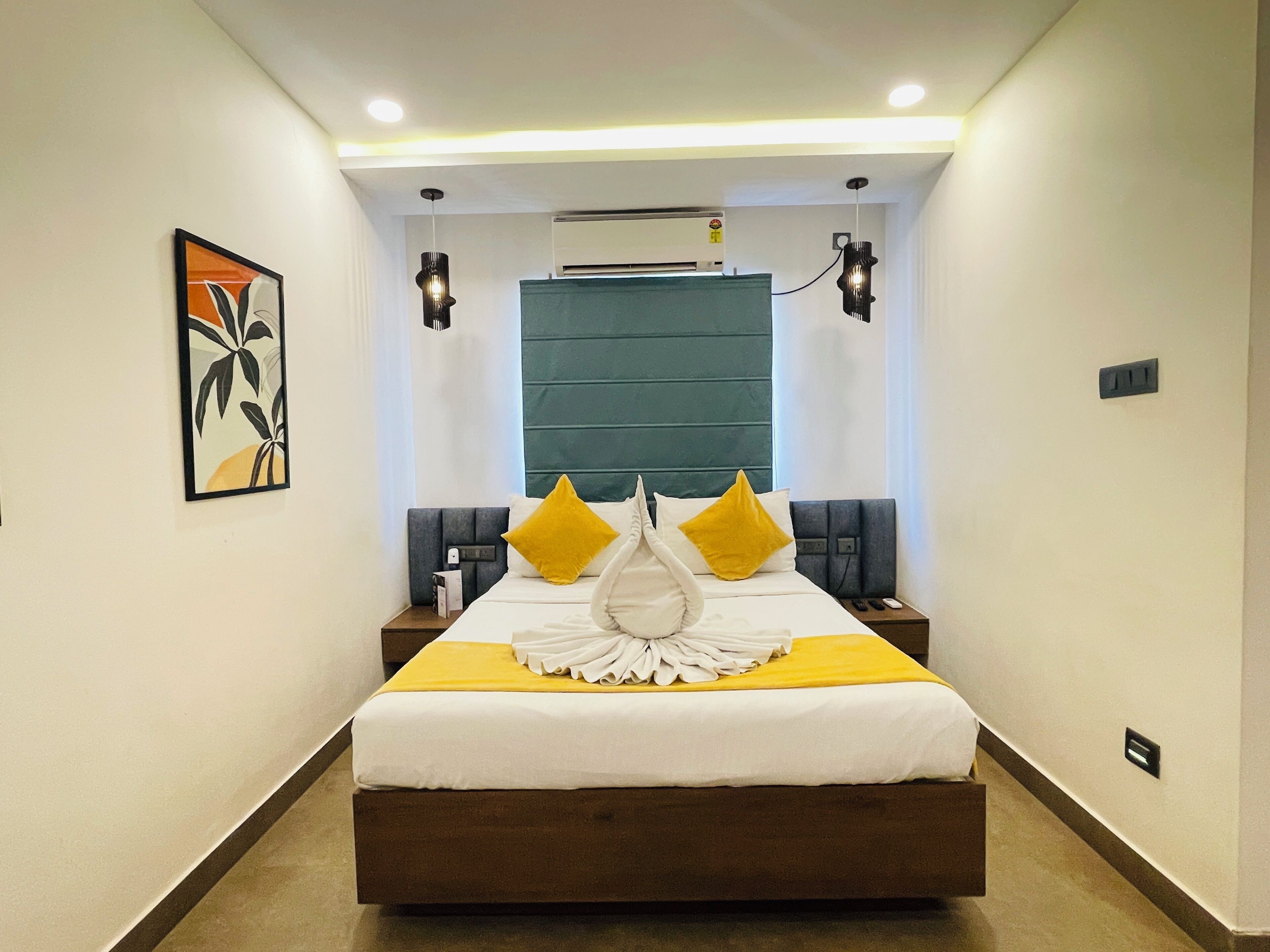 A modern hotel room with a large bed, bedside tables, a wall-mounted air conditioner, and a ceiling light fixture.