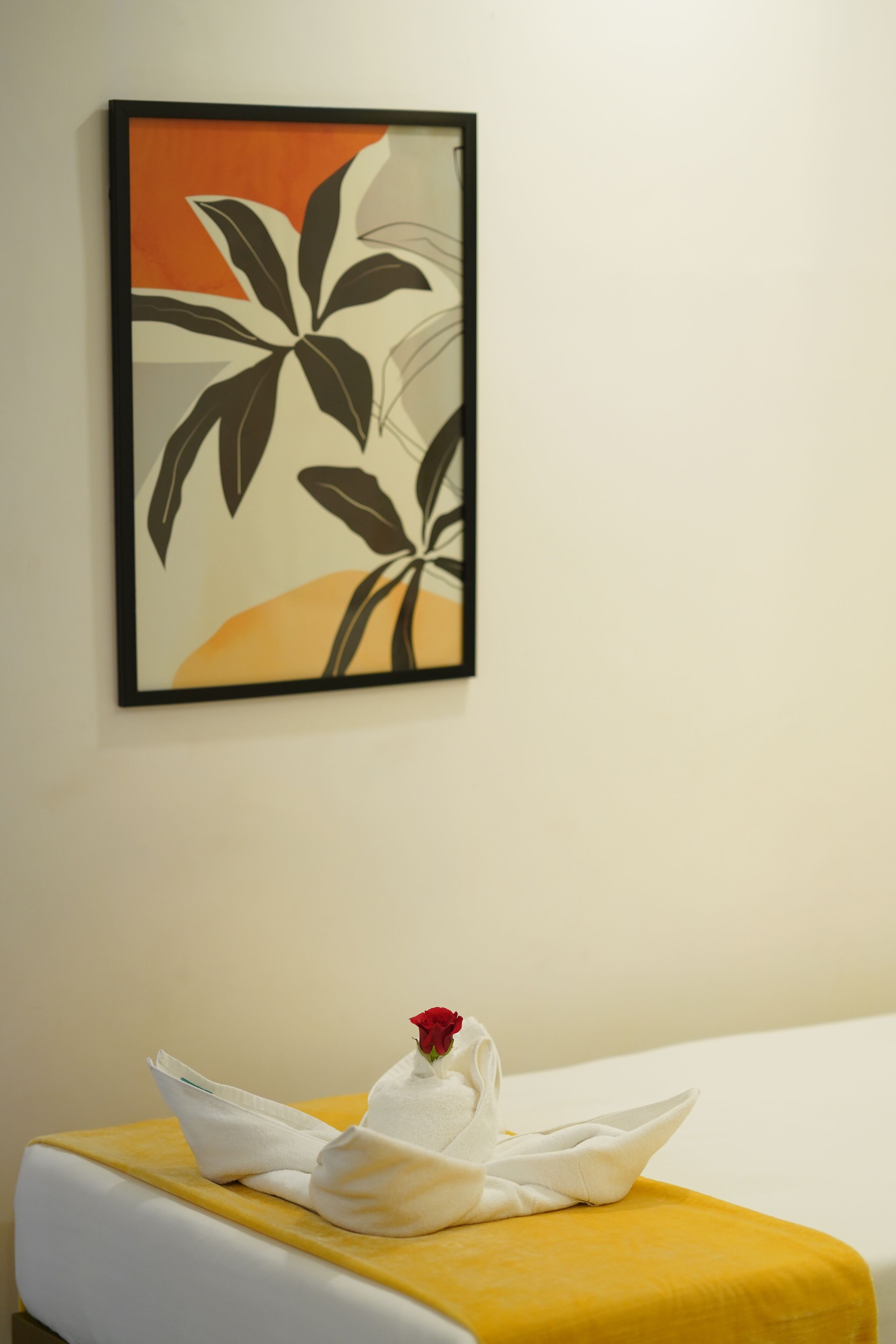 Habitación de hotel con cama, colcha amarilla, toalla blanca con una rosa roja y una obra de arte enmarcada en la pared.