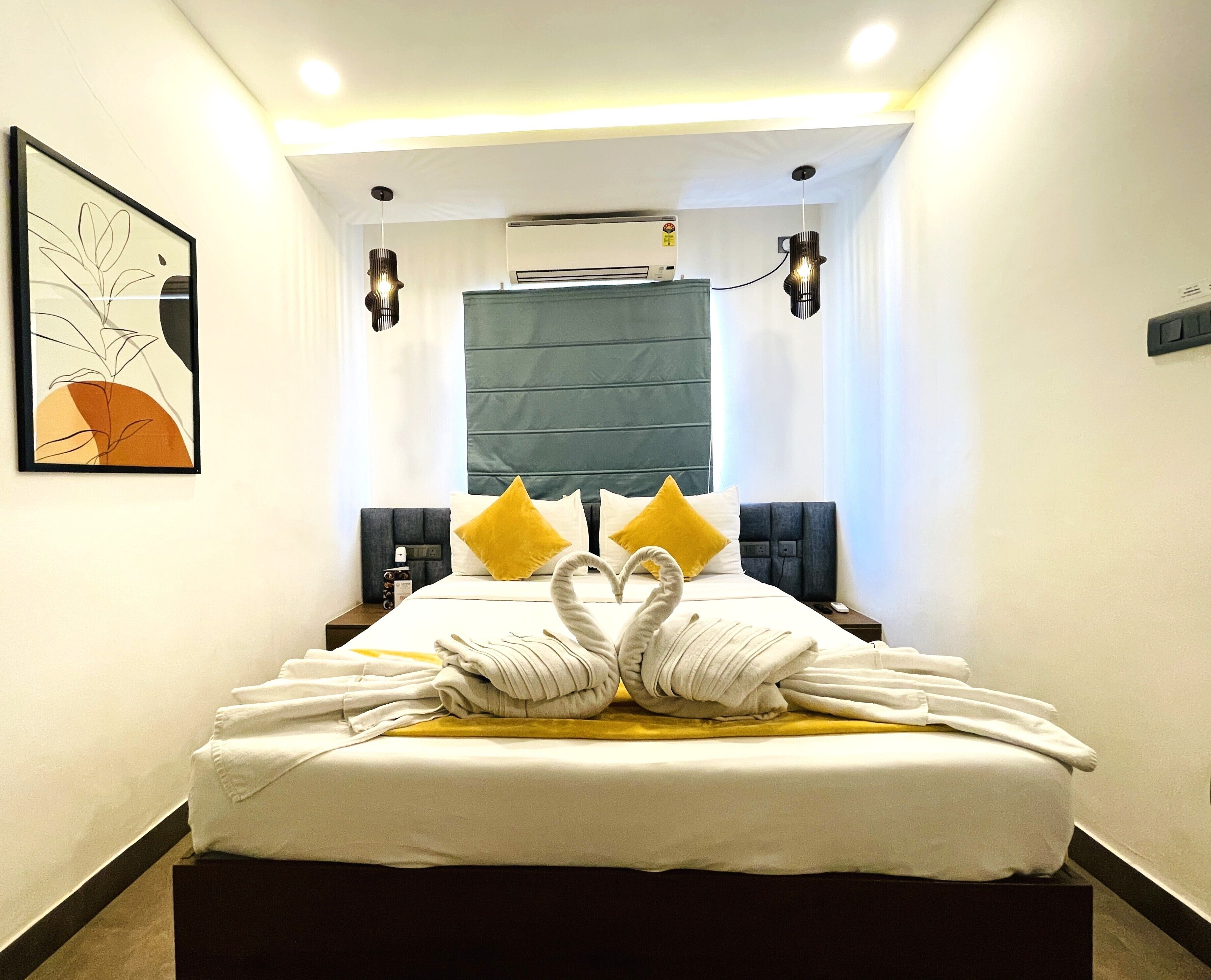 A hotel room with a bed, a wall-mounted air conditioner, a framed artwork, and a ceiling light fixture.