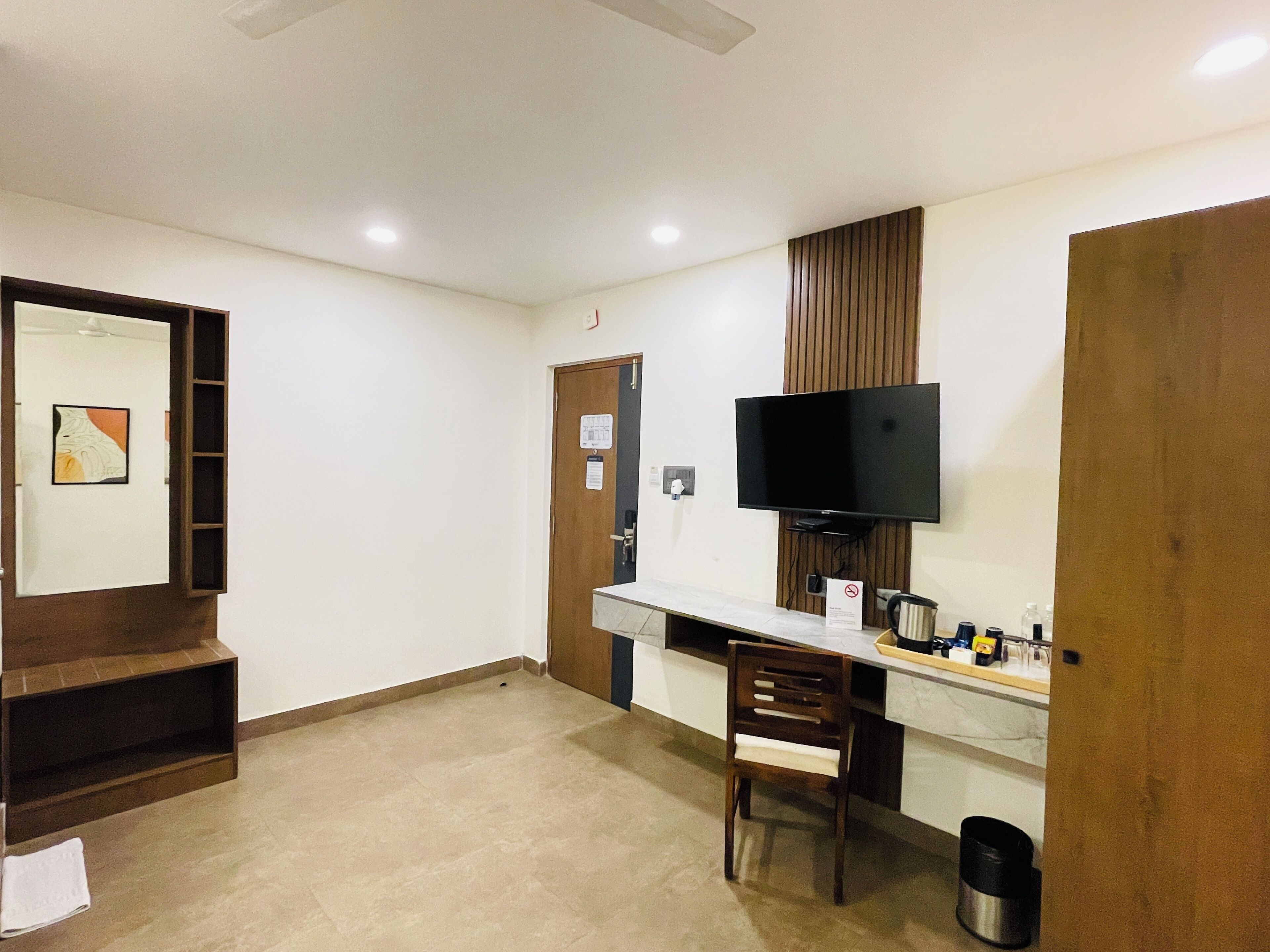 A hotel room with a wooden desk, a television mounted on the wall, a chair, and a small trash bin.