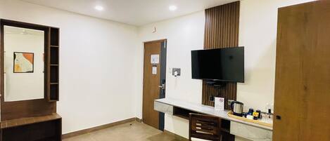 A hotel room with a wooden desk, a television mounted on the wall, a chair, and a small trash bin.