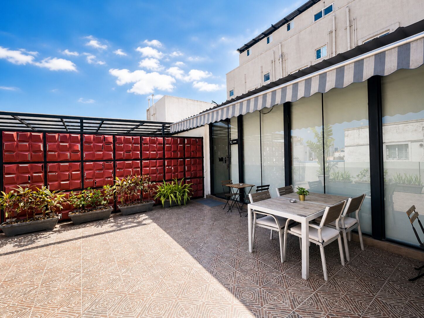 Un patio con suelo estampado, una mesa y sillas, plantas en macetas y una pared roja con motivos geométricos.