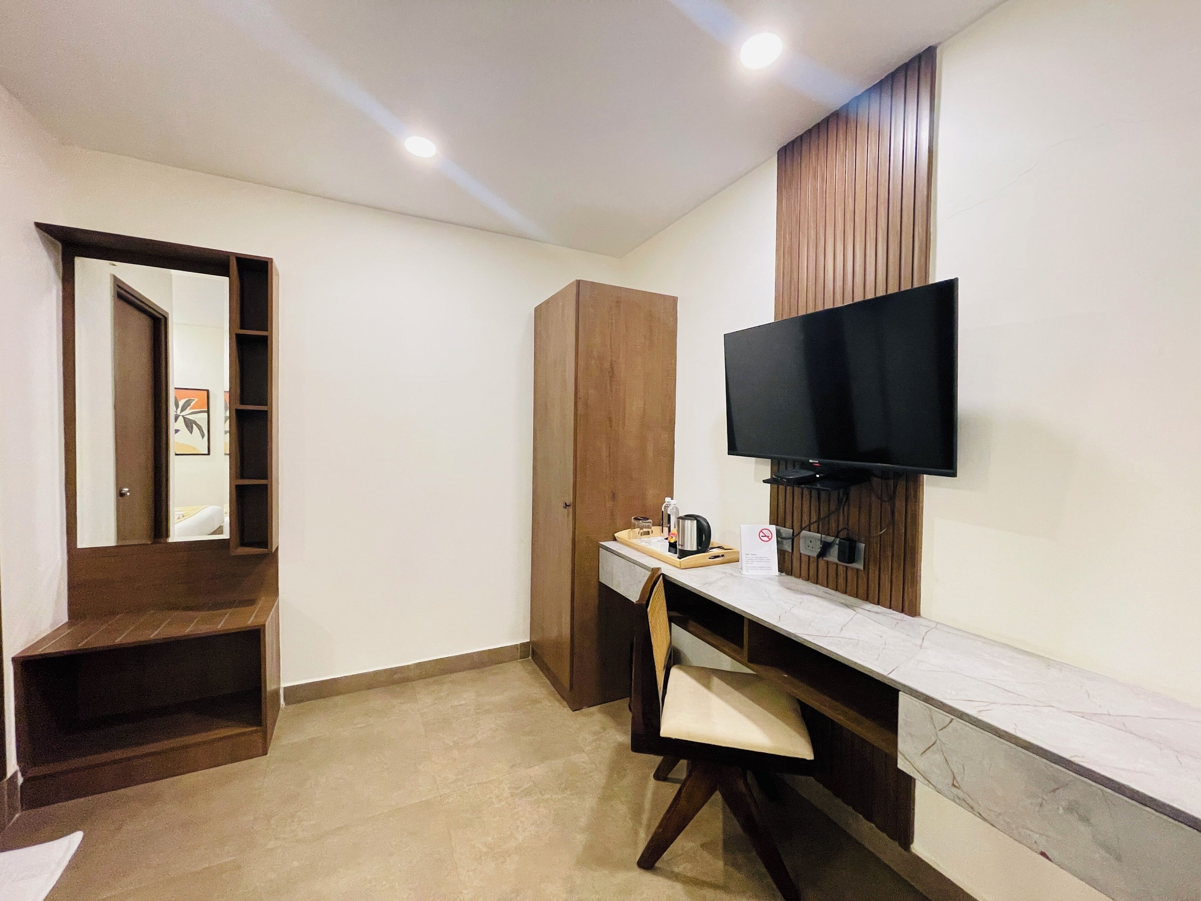 A hotel room with a wooden wardrobe, a flat-screen TV mounted on the wall, a desk with a chair, and a small table with a coffee maker.