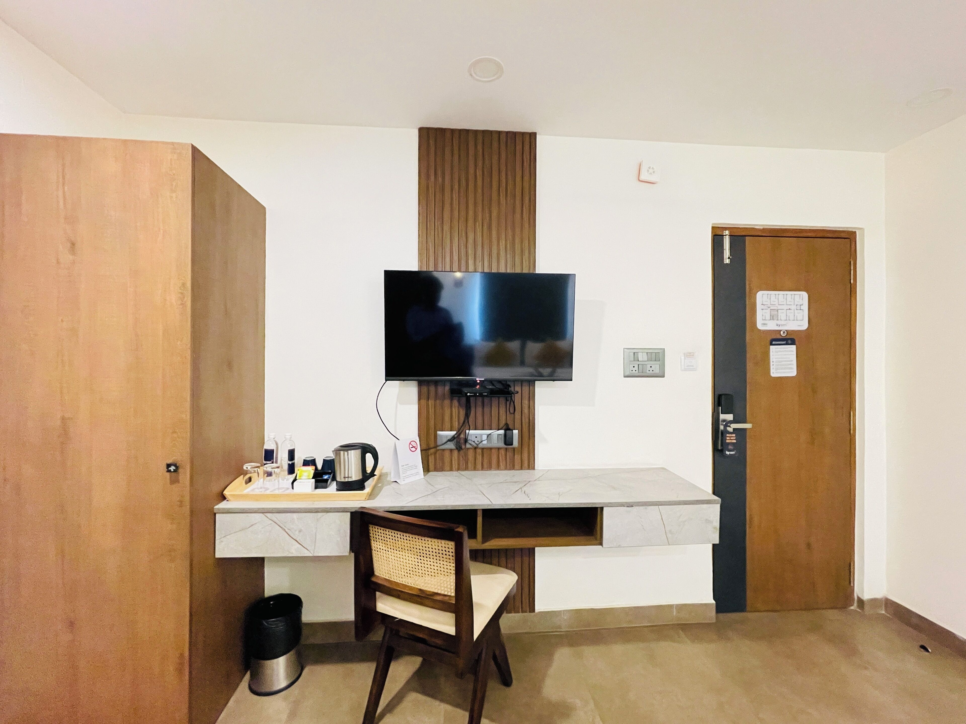 Une petite pièce avec un téléviseur fixé au mur, un bureau avec une chaise, une machine à café et une poubelle.