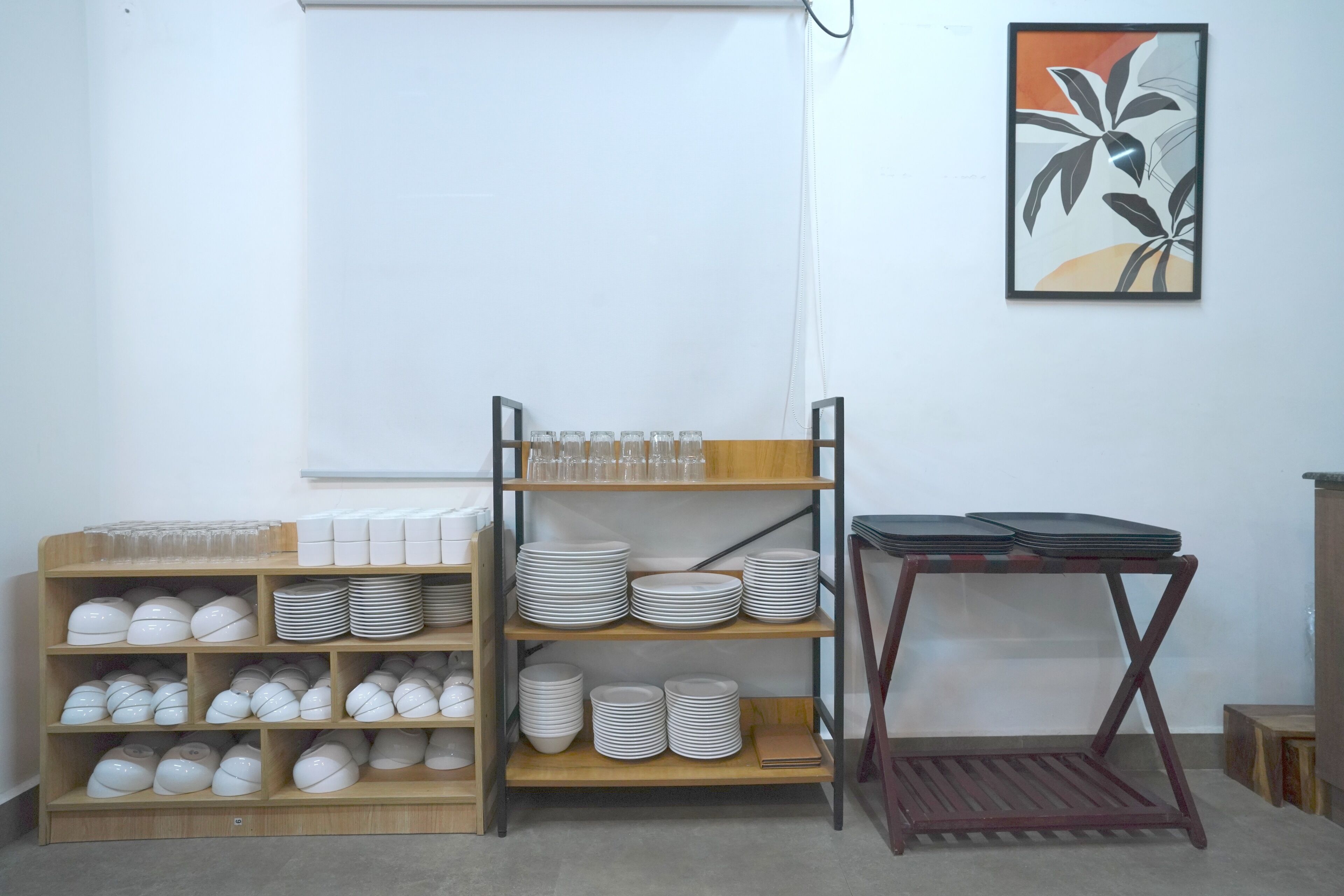 Une pièce avec une étagère en bois où sont exposées des assiettes et des bols, une petite table avec un plateau, et une œuvre d’art encadrée accrochée au mur.