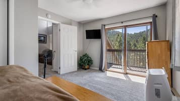 A bedroom with a bed, a desk, a fan, a TV, a sliding glass door to a balcony, and a potted plant.