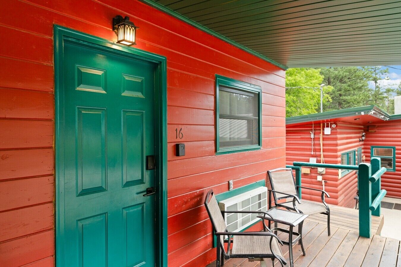 A red wooden building with a green door and window, numbered 16, featuring a porch with two chairs and a small table.