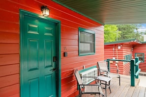 A red wooden building with a green door and window, numbered 16, featuring a porch with two chairs and a small table. - Room 17 – Queen Beds (Lead)
