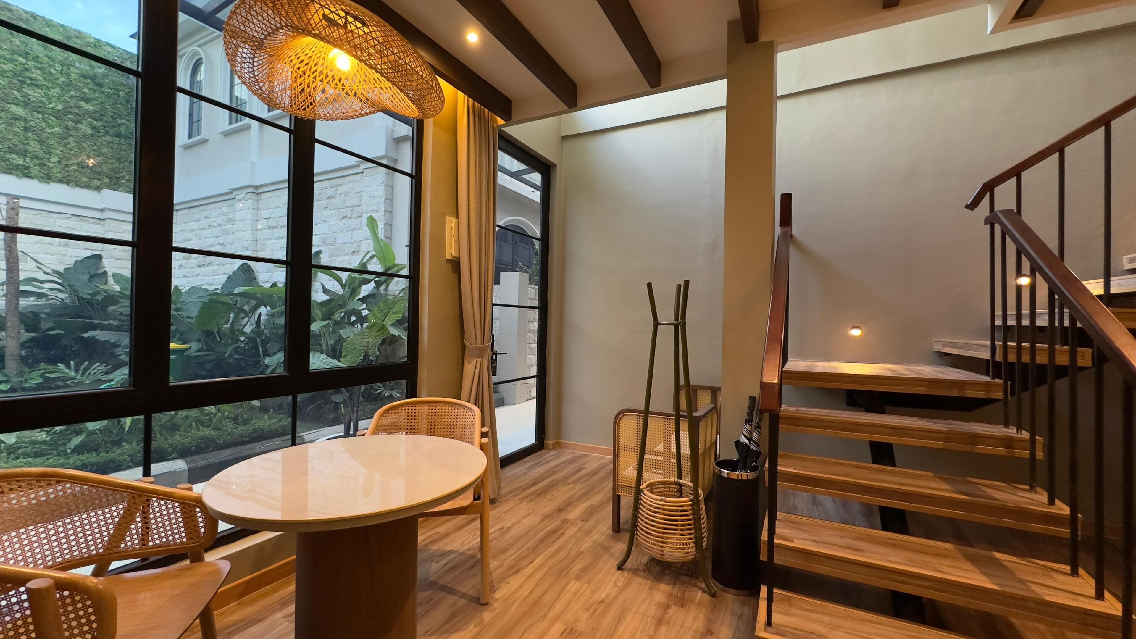 A modern interior with a round table, wicker chairs, a staircase, and large windows overlooking greenery.