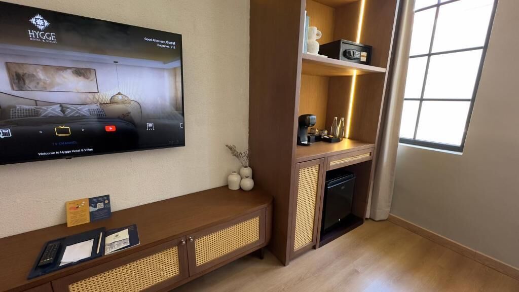 A modern hotel room with a flat-screen TV displaying the hotel's name, a wooden console table with a basket, a small shelf with decorative items, and a window with natural light.