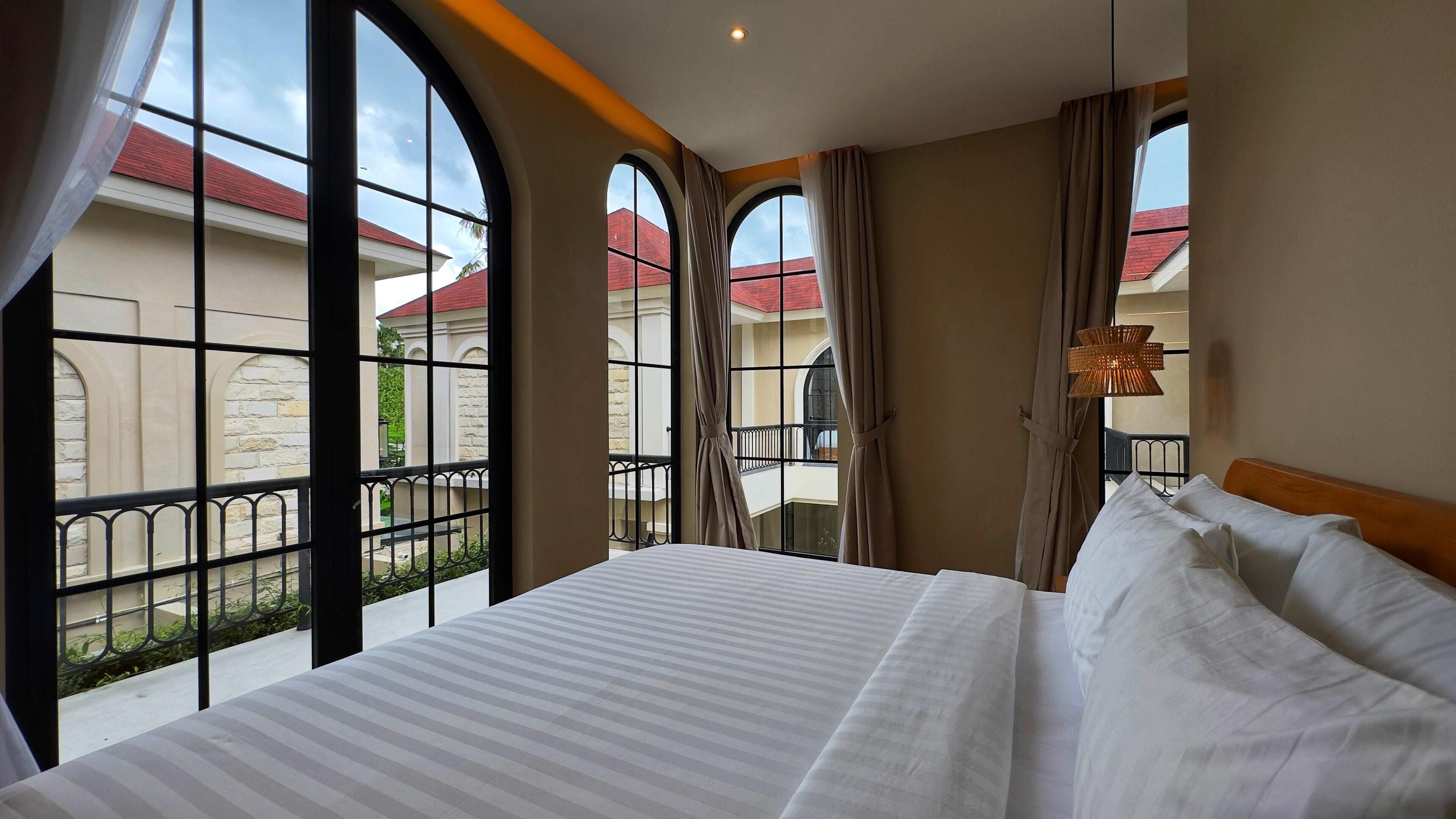 Ein Hotelzimmer mit einem großen Bett, zwei Fenstern mit Vorhängen, einem Balkon mit Geländer und Blick auf Gebäude mit roten Dächern.
