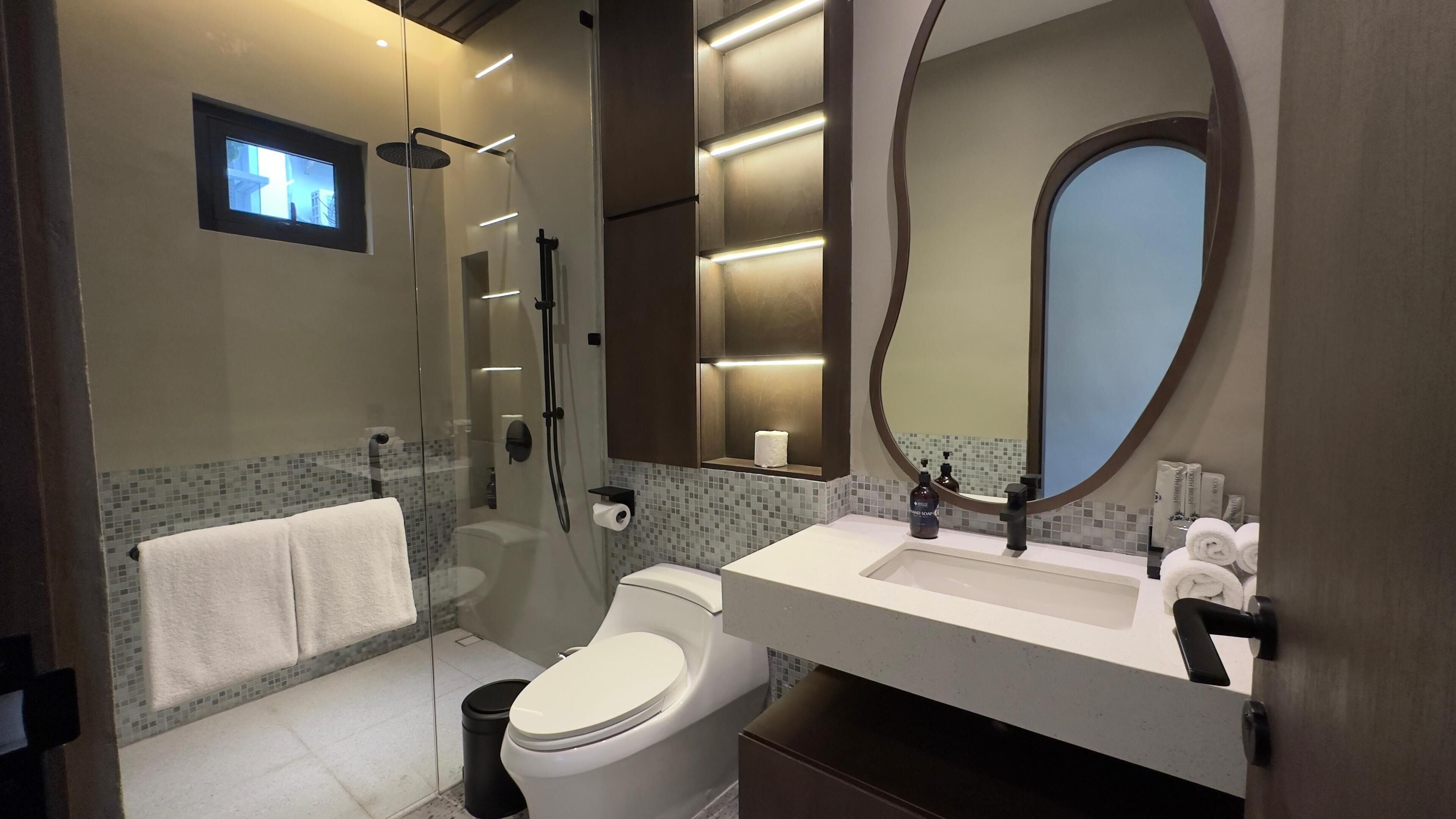 A modern bathroom with a glass-enclosed shower, a toilet, a sink with toiletries, and a large mirror.