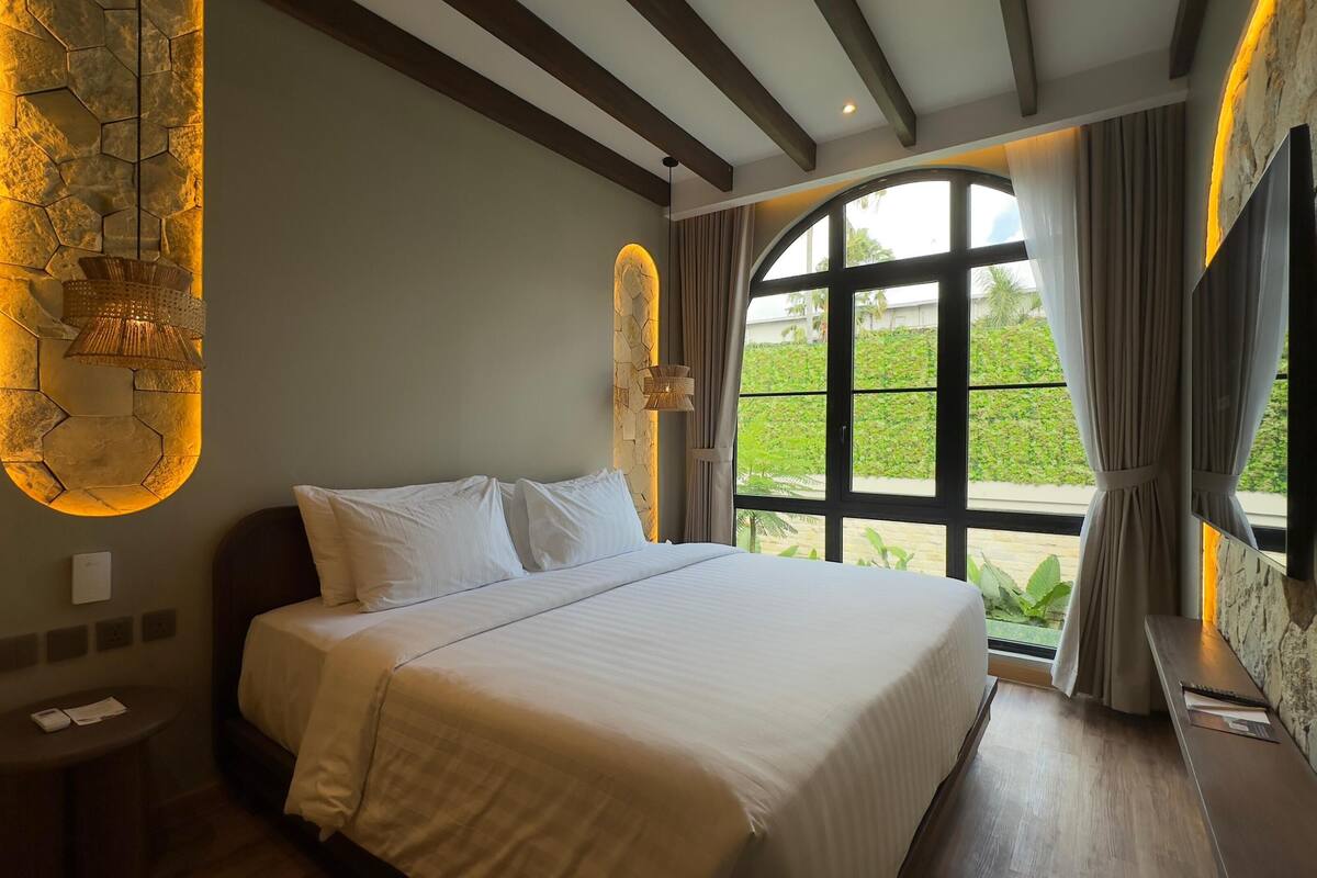 Une chambre avec un grand lit, un mur en pierre servant d’élément décoratif, une fenêtre avec des rideaux et une vue sur la verdure à l’extérieur.