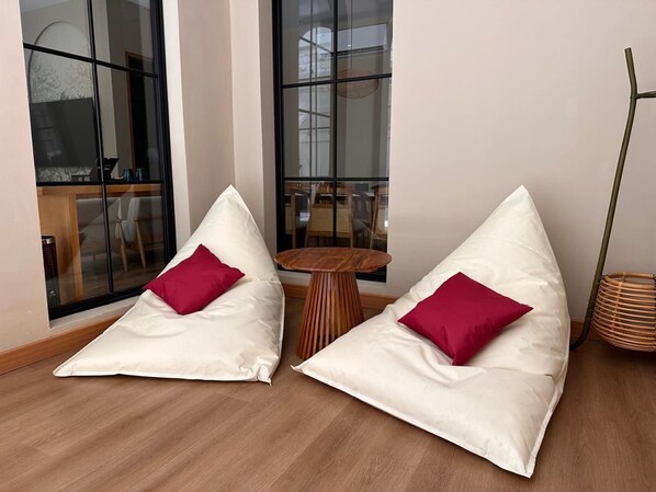 Deux poufs blancs, accompagnés de coussins rouges, se trouvent dans une pièce dont le sol est recouvert de planches de bois. On y aperçoit également une petite table en bois et un panier en osier.