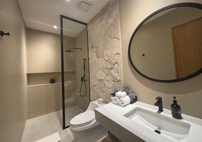 A modern bathroom with a stone wall, a glass-enclosed shower, a white sink with a large mirror, and a toilet.