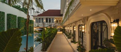 A hotel corridor with a pool, surrounded by lush greenery and architectural details.