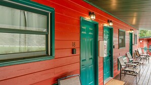 A row of teal doors with a number '18' on a red building facade, outdoor seating with chairs and a bench, and a covered porch area. - Room 16 – Classic Comfort (Lead)