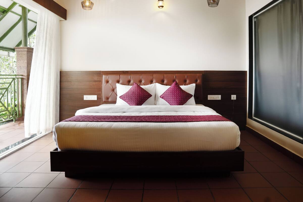 Uma cama bem arrumada com cabeceira de madeira, almofadas brancas e roxas, e um cobertor vermelho.